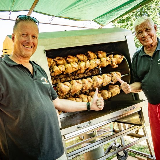 Zwei Männer stehen neben einem Hähnchen-Drehspieß, beide zeigen den Daumen nach oben. Der Mann auf der linken Seite hat eine Sonnenbrille auf dem Kopf.