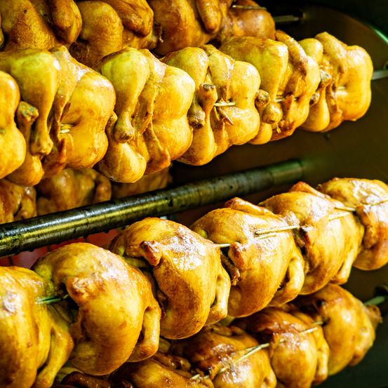 Mehrere ganze Hähnchen werden am Spiess in einem Ofen geröstet. Die Hähnchen haben knusprige goldene Haut und scheinen vollständig gegart zu sein.