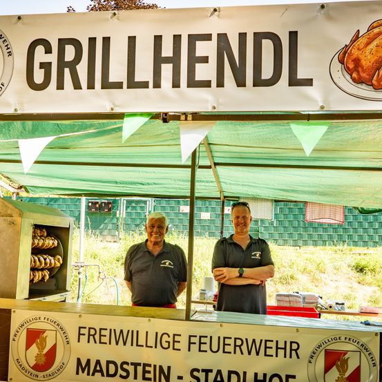 Zwei Männer stehen hinter einem Tresen an einem Imbissstand. Ein Banner zeigt 'GRILLHENDL'. Die Männer tragen Uniformen einer Feuerwehr.