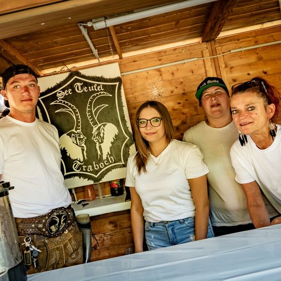 Vier Personen posieren vor einer hölzernen Kabine mit einem Banner, das 'See Teufel' und ein Widder-Emblem zeigt. Einer trägt Hut und Gürtel, während zwei Brillen und weiße T-Shirts tragen. Eine Frau lächelt.