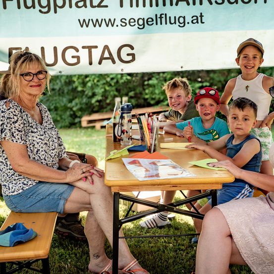 Eine Frau und vier Kinder sitzen an einem Picknicktisch im Freien, lächeln und betrachten ein Blatt Papier. Dahinter steht ein Banner mit der Aufschrift 'Flugtag'.