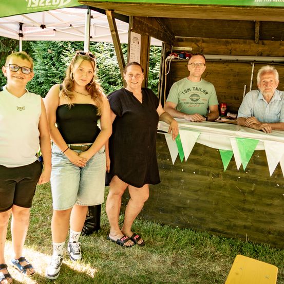 Fünf Menschen stehen vor einem hölzernen Stand. Der Stand hat ein weißes Dach, und eine grüne Fahne hängt an der Vorderseite. Drei Frauen stehen und zwei Männer sitzen am Tresen.