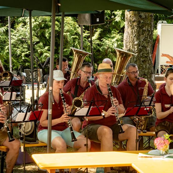 Eine Band probt im Freien unter einem Regenschirm. Sie spielen verschiedene Instrumente, darunter Saxophone, Trompeten und Klarinetten. Die Musiker sitzen auf Bänken mit Notenständern.