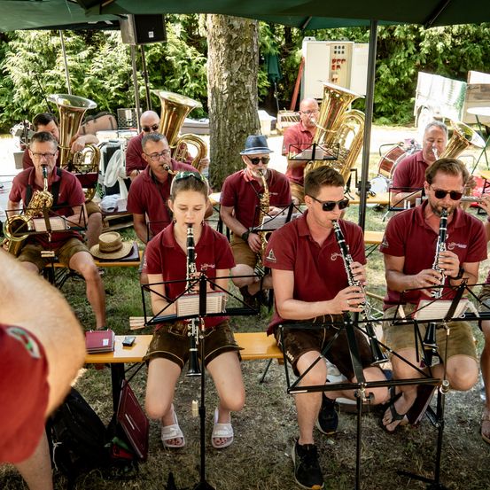 Eine Band übt draußen unter einem grünen Zelt, mit verschiedenen Instrumenten wie Tuba, Saxophon und Klarinette. Sie tragen burgunderrote Hemden und Shorts und sitzen auf Holzbänken.