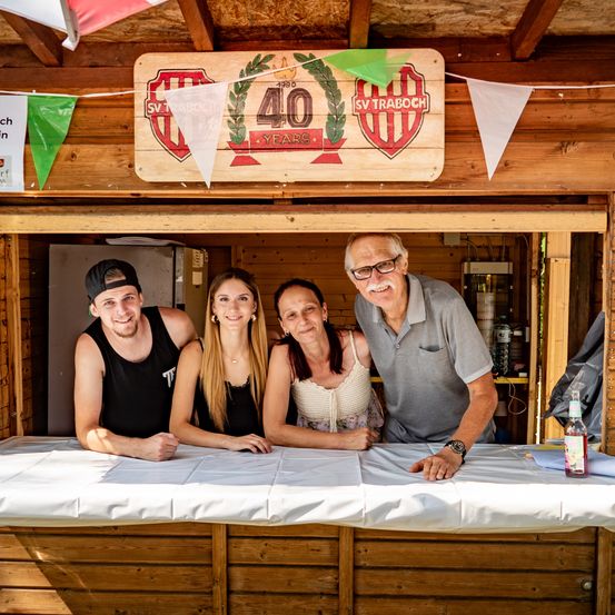 Vier Menschen lächeln in einer hölzernen Kabine mit einem Schild, das '40 Jahre' zeigt. Ein Mann und zwei Frauen tragen ärmellose Shirts, und der Mann trägt eine Kappe.