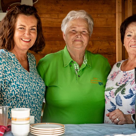 Drei Frauen stehen hinter einem Tisch mit Tassen und Tellern und lächeln. Eine trägt ein grünes Poloshirt, eine andere ein Blumenkleid und die dritte ein blaues Oberteil. Hinter ihnen befinden sich Holzwände.