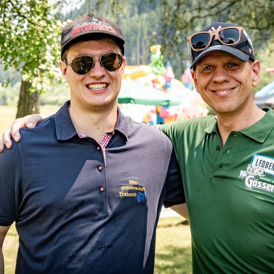 Zwei lächelnde Männer, einer in einem blauen Shirt, der andere in Grün, beide mit Mützen und Sonnenbrillen, stehen in einem Grasbereich. Dahinter befindet sich ein farbenfroher aufblasbarer Spielbereich.