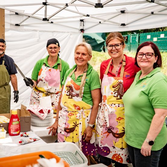 Fünf Frauen in grünen Hemden und Schürzen stehen unter einem Zelt und lächeln für ein Foto. Ein Mann steht hinter ihnen mit einer Grillzange. Verschiedene Gegenstände stehen auf dem Tisch, einschließlich Teller, einer Flasche und einer Schachtel.