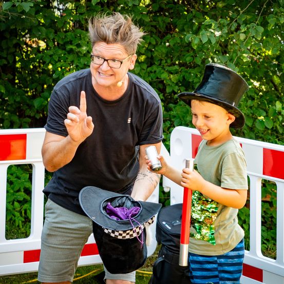 Ein Mann und ein Junge lächeln und posieren für ein Foto. Der Mann hält einen Hut und der Junge hält einen Zauberstab. Sie tragen beide Hüte und stehen vor einem weißen Zaun.