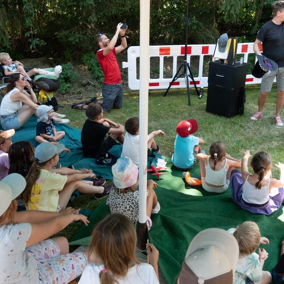 Ein Mann macht ein Foto von Kindern, die auf einer Decke im Park sitzen. In der Nähe steht ein Mann und ein schwarzer Kasten liegt auf dem Rasen.