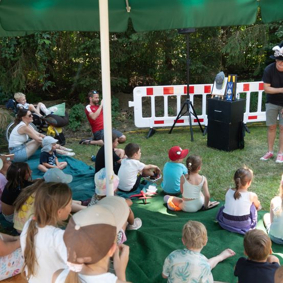 Eine Gruppe von Kindern und Erwachsenen sitzt auf einer Decke unter einem Schirm in einem Park. Ein Mann spricht in ein Mikrofon.