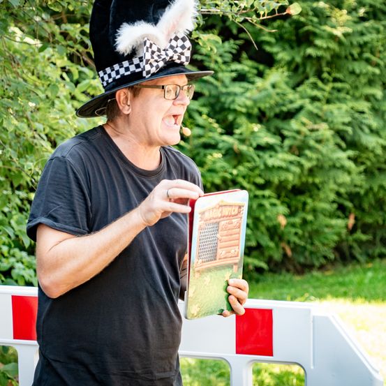 Ein Mann mit Zylinderhut und Brille liest ein Buch laut vor sich hin, vermutlich bei einer Veranstaltung.