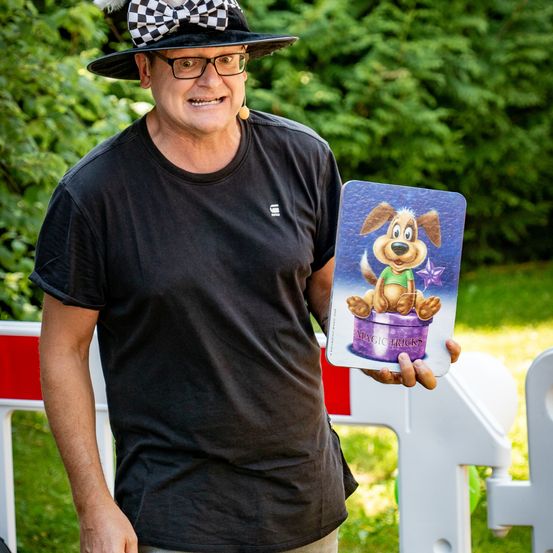 Ein Mann in einem schwarzen T-Shirt und einem karierten Hut hält eine Karte mit einem gezeichneten Hund in einem grünen Hemd. Er scheint einen Zaubertrick durchzuführen.