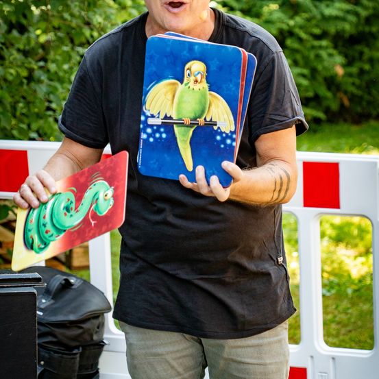 Ein Mann in einem schwarzen T-Shirt und Jeans hält einen Stapel bunter Karten in der Hand, eine zeigt einen Papagei und die andere eine Schlange, in einer Außenumgebung.