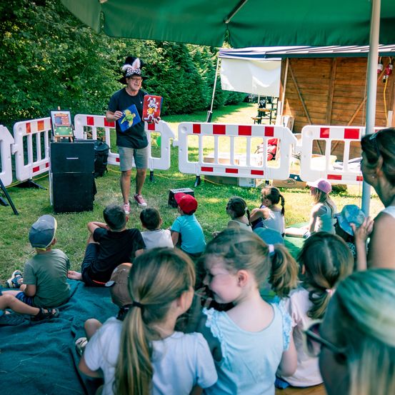 Ein Mann in einem Hut steht unter einem Zelt und liest einem Kreis von Kindern, die auf dem Gras sitzen, ein Buch vor.
