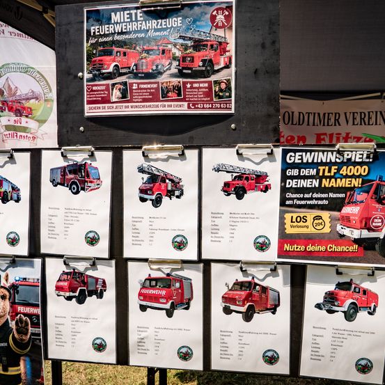Ein Anzeigebrett mit einem Plakat, das Feuerwehrfahrzeuge für besondere Momente anbietet. Unten sind mehrere Feuerwehrfahrzeuge mit ihren Spezifikationen und Bildern ausgestellt. Ein weiteres Plakat bietet die Chance, einen Namen auf dem Feuerwehrfahrzeug zu gewinnen.