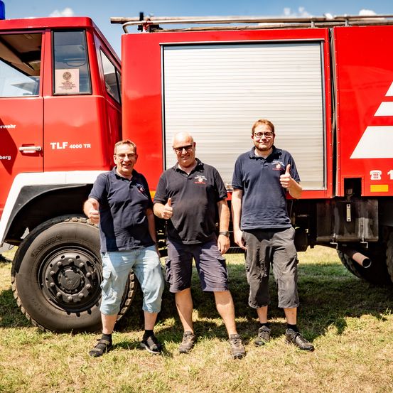 Drei Männer stehen vor einem Feuerwehrauto. Der Mann in der Mitte zeigt Daumen hoch, und alle lächeln. Das Auto hat ein Schild mit der Aufschrift TLF 4000 TR.