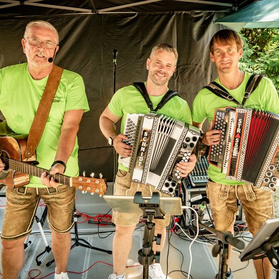 Drei Männer in grünen Shirts und Shorts spielen auf einer Bühne Musikinstrumente, einer spielt Gitarre und die anderen spielen Akkordeons.