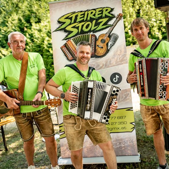 Drei Männer in grünen Shirts und braunen Shorts stehen draußen. Der Mann in der Mitte hält ein Akkordeon, der linke Mann hält eine Gitarre und der rechte Mann hält ein anderes Akkordeon. Sie stehen vor einem Poster.
