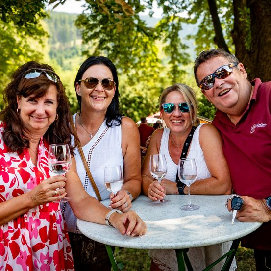 Fünf Menschen sitzen um einen Tisch in einem Park, alle mit Sonnenbrille und lächeln. Sie halten Weingläser, und eine Frau links trägt ein rotes Blumenkleid.