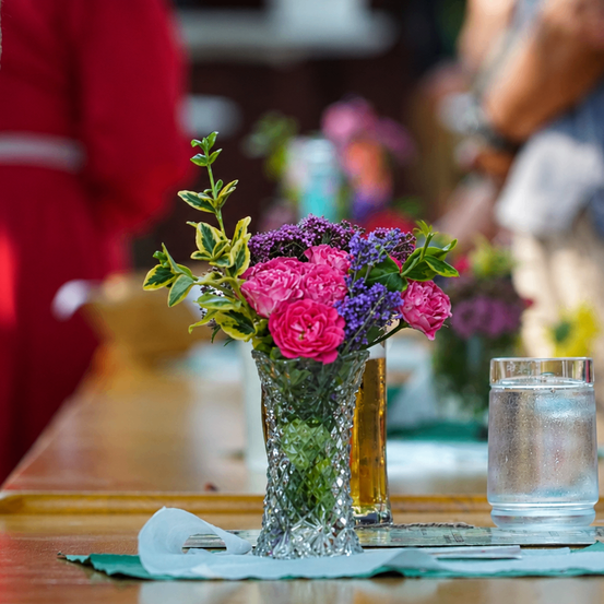 Ein hölzerner Tisch mit einer Glasschale, die einen Strauß aus rosa und lila Blumen hält. Es steht auch ein Glas Wasser auf dem Tisch.