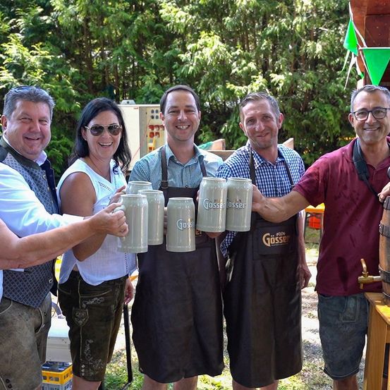 Fünf Erwachsene halten Bierkrüge bei einer Veranstaltung im Freien. Drei Männer in Schürzen, eine Frau mit Sonnenbrille und ein Mann in einem Vest. Bäume und ein grüner Banner im Hintergrund.