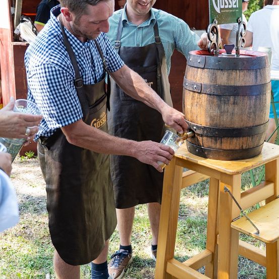 Zwei Männer in Schürzen arbeiten auf einem Festival an einem Holzfass, einer gießt ein Getränk in ein Glas.