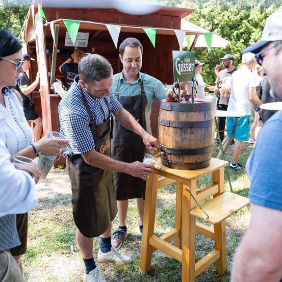 Zwei Männer in Schürzen zapfen Bier aus einem Fass bei einer Veranstaltung im Freien. Eine Menschenmenge beobachtet, wie ein Mann das Fass zapft. Ein Stand mit Wimpelgirlanden im Hintergrund.