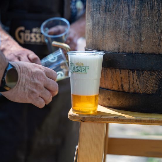 Eine Person schenkt Bier aus einem Fass in ein Glas auf einem Holzständer. Das Glas hat das Logo von Gosset.