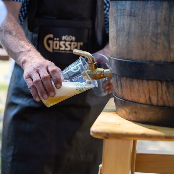 Ein Mann schenkt Bier aus einem Fass in ein Glas auf einem Holztisch. Das Fass hat einen Messinghahn.