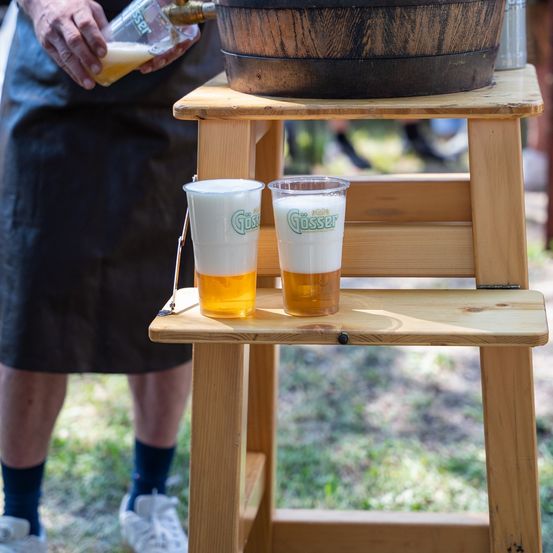 Eine Person gießt Bier aus einer Flasche in zwei Gläser auf einem Holzständer, mit einem Fass darüber. Die Gläser haben das Wort Goesser darauf.