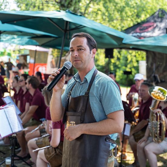 Ein Mann mit einer Lederschürze spricht in ein Mikrofon in einem Außenbereich, hinter ihm befindet sich ein Orchester.