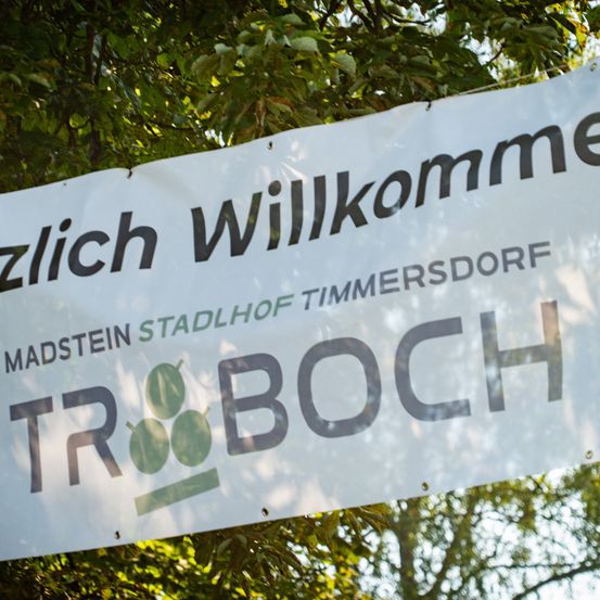 Ein weißes Banner mit schwarzem Text, das an einem Baum hängt, zeigt die Aufschrift 'TR BOOCH' und 'Madstein stadlhof Timmersdorf'. Das Banner wirbt wahrscheinlich für eine lokale Veranstaltung oder ein Unternehmen.