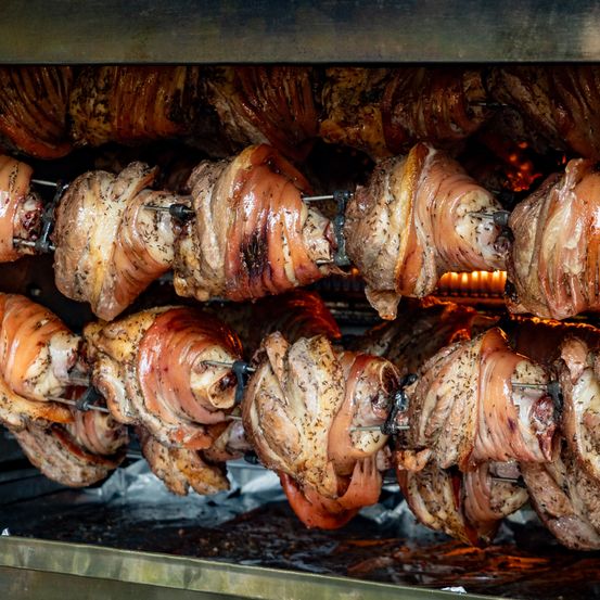 Ein Nahaufnahme-Blick auf verschiedene Hähnchenschenkel, die in einem Ofen geröstet werden. Sie sind mit Kräutern und Gewürzen verfeinert und einige sind in Speck gewickelt. Die Hitze des Ofens verleiht ihnen ein knuspriges und goldbraunes Aussehen.