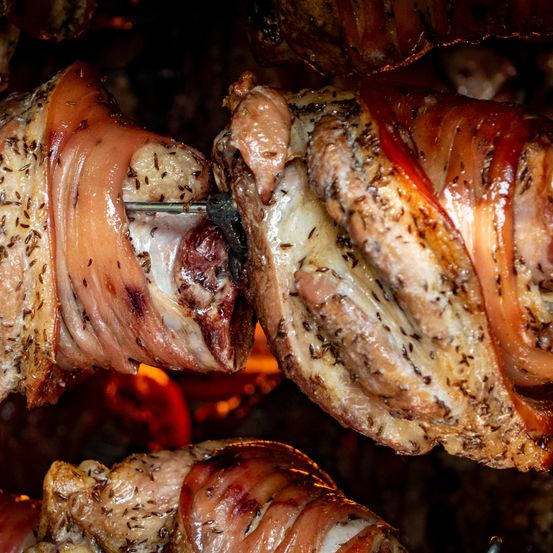 Nahaufnahme von gekochtem Fleisch, das in Speck eingewickelt und mit Kräutern gewürzt ist, wird über offenem Feuer gegrillt. Das Fleisch ist verkohlt und hat eine knusprige Textur.