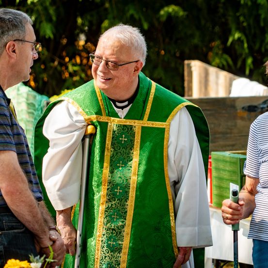 Ein älterer Priester in grünen und weißen Roben lächelt und hält einen Gehstock. Er wird von Menschen umgeben, einer hält Blumen, in einem Garten.