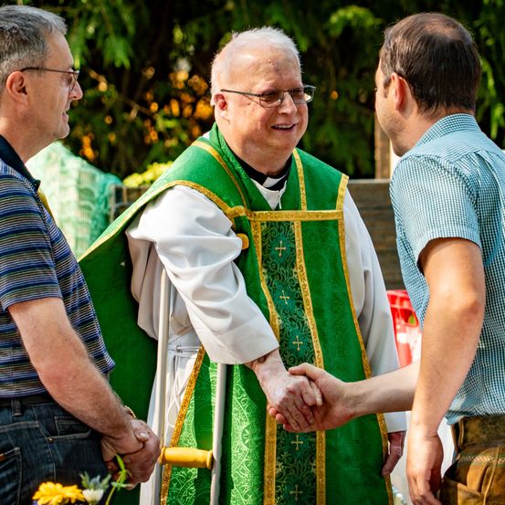 Ein Priester mit einer grünen und weißen Robe schüttelt die Hände von zwei Männern, von denen einer eine Brille trägt und der andere ein blaues Hemd, in einem Garten.