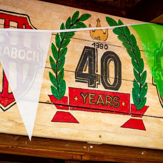 Ein Holzschild mit einem Lorbeerkranz und der Nummer 40, was auf ein 40-jähriges Jubiläum hinweist. Links hängt eine weiße Fahne.