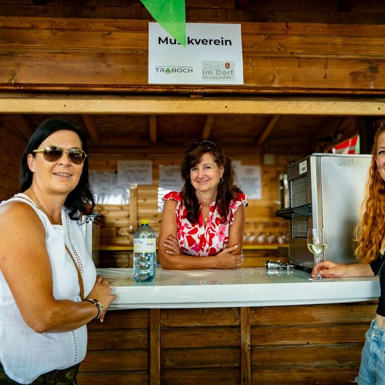 Drei Frauen stehen hinter einer Theke in einer hölzernen Bar. Eine Wasserflasche und ein Weinglas stehen auf der Theke. Die Frau rechts hält ein Weinglas.
