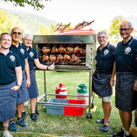 Fünf Männer in Schürzen stehen neben einem Bratenofen mit Fleisch im Freien. Sie lächeln und schauen in die Kamera. Hinter ihnen ist ein Feuerwehrauto zu sehen.
