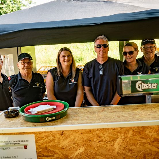 Fünf Personen lächeln und posieren für ein Foto an einem Bierstand. Ein grüner Eimer mit dem Wort Gösser steht auf der Theke. Der Mann links trägt einen Hut.