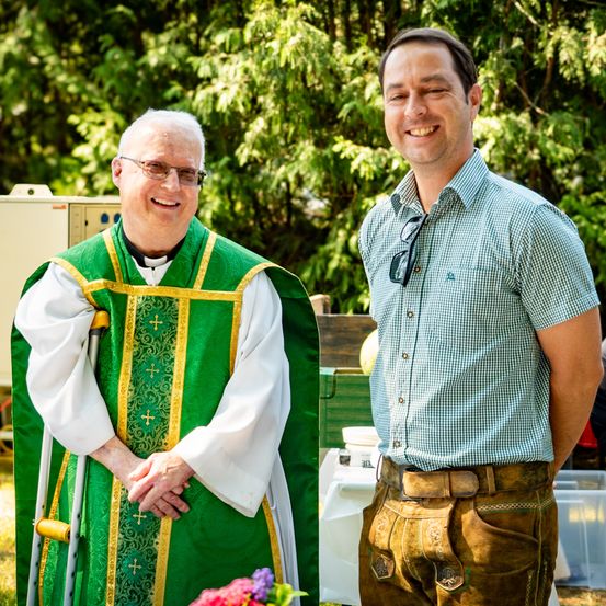 Ein Priester mit einem Gehstock und ein lächelnder Mann in einem grün-weiß karierten Hemd. Sie stehen draußen in der Nähe eines Tisches mit Blumen und Bäumen im Hintergrund.