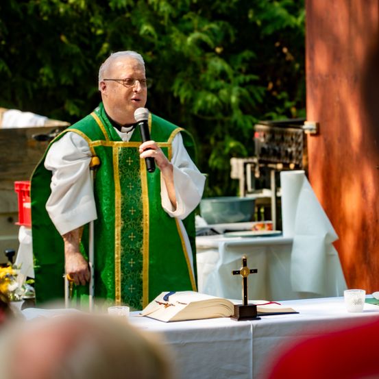 Ein Priester in einer grünen Robe steht hinter einem Tisch, hält ein Mikrofon und spricht zu einer Gruppe von Menschen. Ein offenes Buch und ein Kreuz liegen auf dem Tisch.