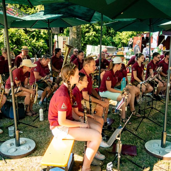 Eine Band sitzt auf einem Rasenbereich unter grünen Regenschirmen und bereitet sich mit ihren Instrumenten auf einen Auftritt vor. Sie tragen burgunderrote Hemden und einige haben Hüte auf. Musikinstrumente sind auf dem Boden verteilt.