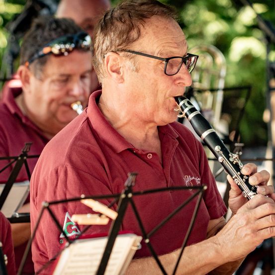 Ein Mann mit Brille und einem weinroten Hemd spielt die Klarinette in einer Freiluftveranstaltung mit anderen Musikern.