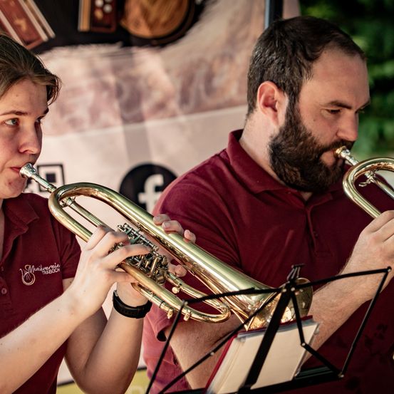 Zwei Musiker in burgunderroten Hemden spielen Trompeten mit Notenständern vor sich, wahrscheinlich bei einer Veranstaltung im Freien.