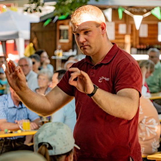 Ein Mann in einem weinroten Poloshirt macht mit den Händen Gesten vor einem Publikum in einem Biergarten.