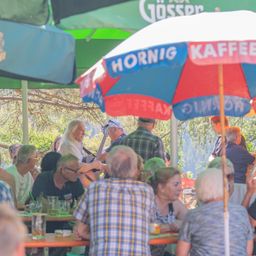 Eine Gruppe von Menschen sitzt unter einem Schirm mit dem Schriftzug 'HORNIG KAFFEE'. Eine Frau spielt Gitarre im Hintergrund.