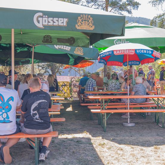 Eine Gruppe von Menschen sitzt unter Schirmen an Tischen in einem Biergarten im Freien. Einige Tische haben Gösser-Bierschirme. Im Hintergrund sind Bäume und Berge zu sehen.