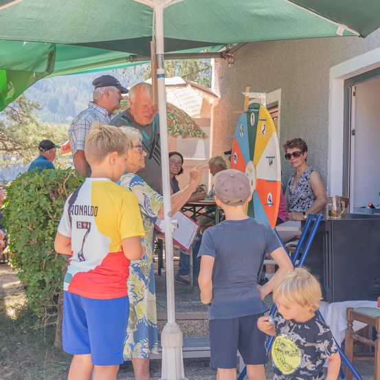 Eine Gruppe von Menschen, einschließlich Kindern, steht unter einem grünen Regenschirm vor einem Haus. Eine Frau hält ein Papier und ein Mann spricht mit ihr. Ein buntes Rad ist an der Wand.